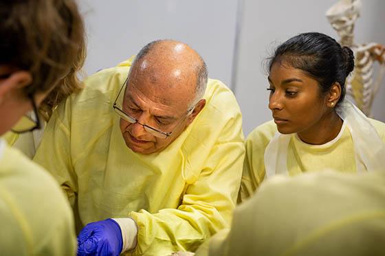 Photo of a medical professional surrounded by students in yellow protective suits and medical caps, perform a medical procedure. 