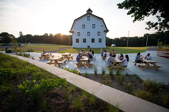 Photo of several people gathering at picnic tables on Chatham University's Eden Hall Campus in front of a white barn. 