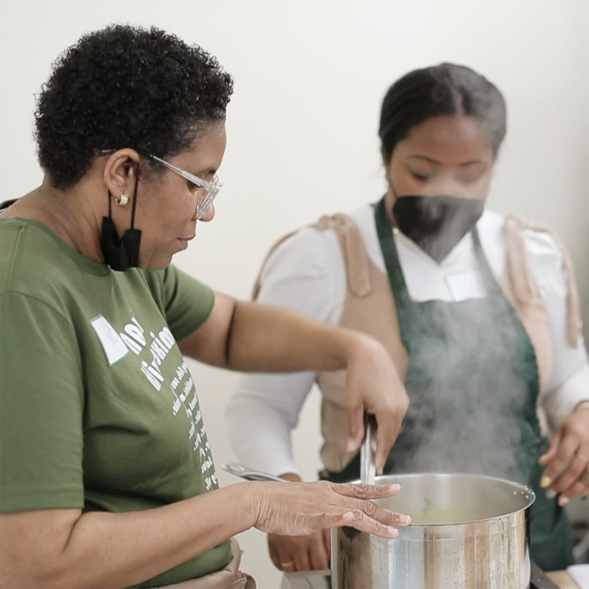 Photo of a Black woman, Toni Simpson, 在锅里搅拌食物，并向一位年轻的黑人妇女示范，她围着围裙，戴着面具，正在观察