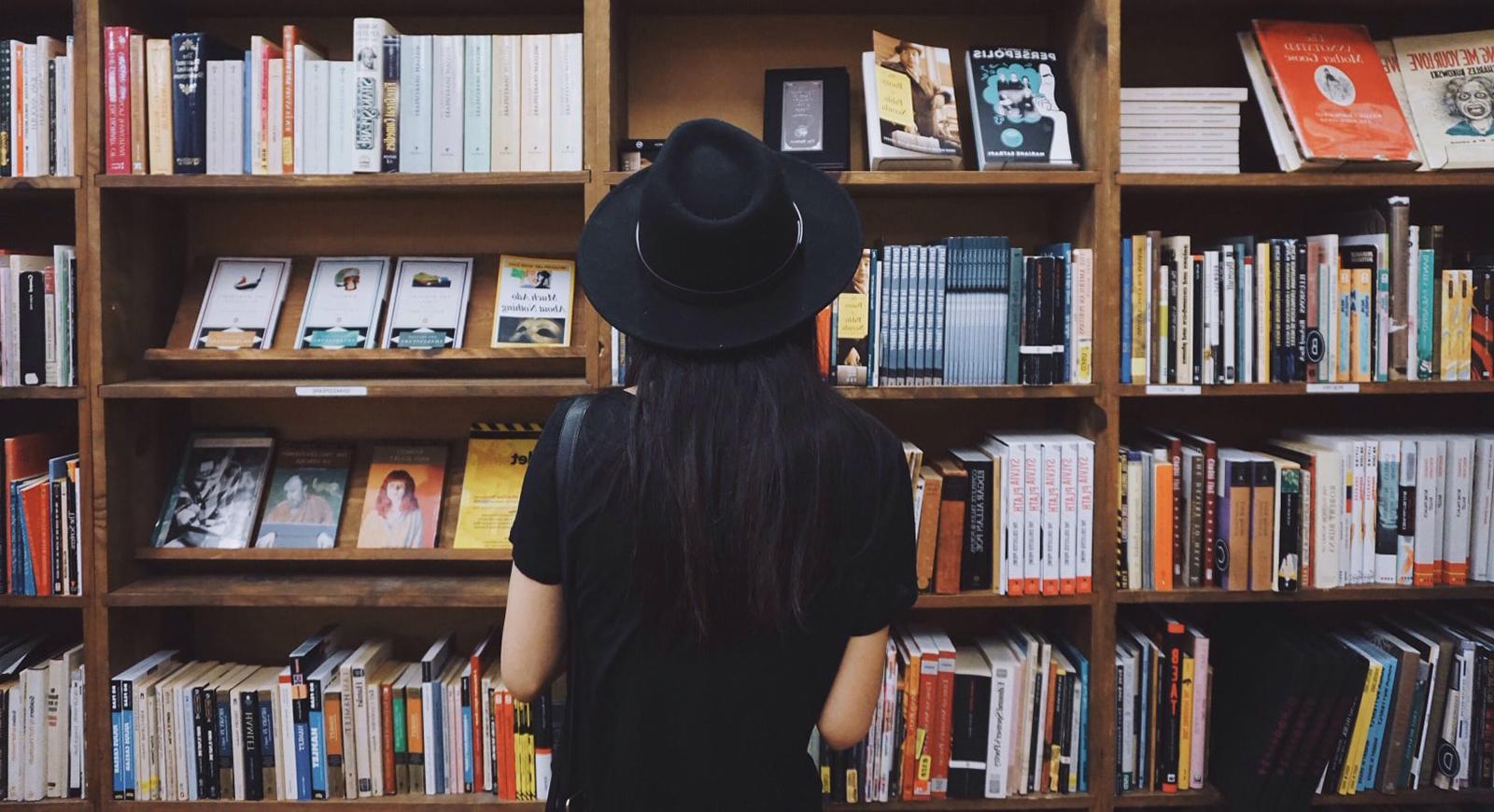Photo of a woman wearing a black wide-brimmed hat and black t-shirt stands in front of a colorful bookshelf. 