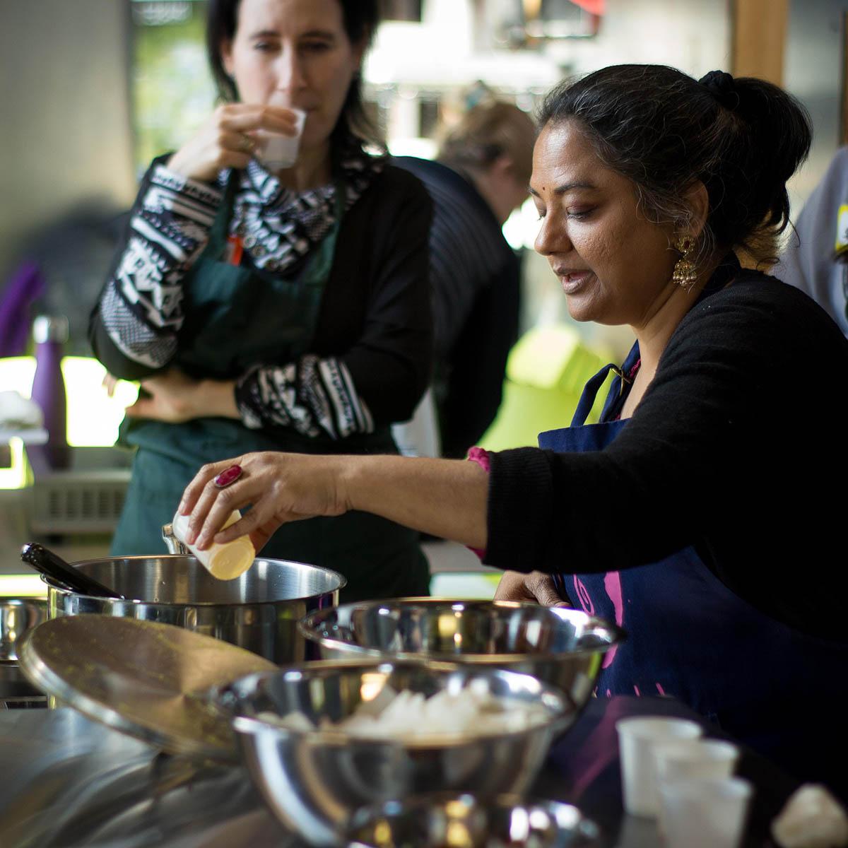 Photo of a South Asian woman demonstrating how to cook, while a class observes