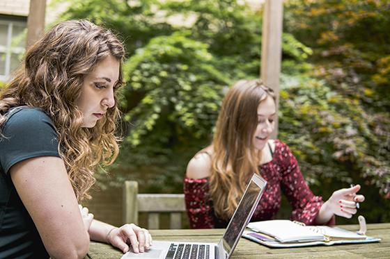 图为两名波胆网站的学生坐在户外野餐桌旁，用笔记本电脑在笔记本上写东西. 