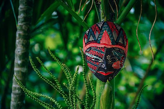 Photo of a colorful mask hanging outside surrounded by greenery. 