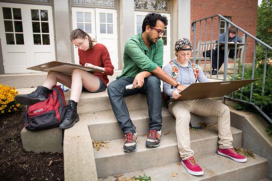 Photo of a Chatham University art professor sitting on steps outside with students who are 画 on art boards. 