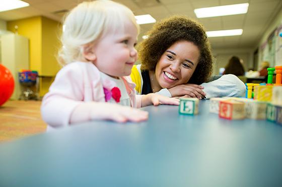 Photo of a smiling Chatham University student leaning over a table smiling at a young toddler who is playing with letter blocks