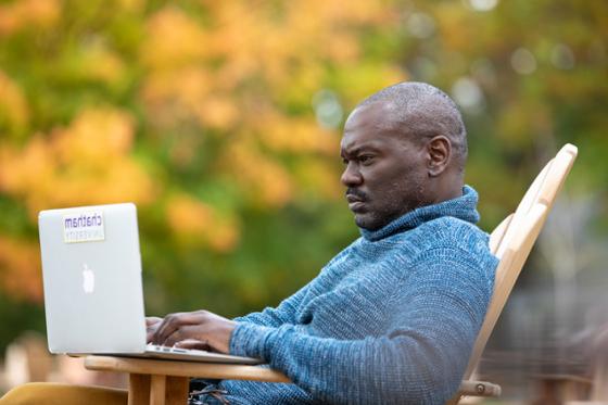 Photo of a Chatham University male student working on his laptop outside, with autumnal leaves in the background.