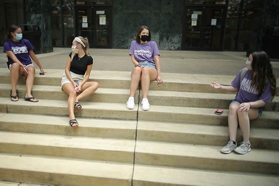 Photo of four female Chatham University students, wearing masks and Chatham t-shirts, all sit socially distant on the library steps