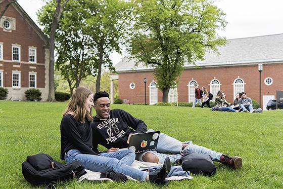 Two Chatham University students sit on a green grass academic quad, working together on a laptop. 