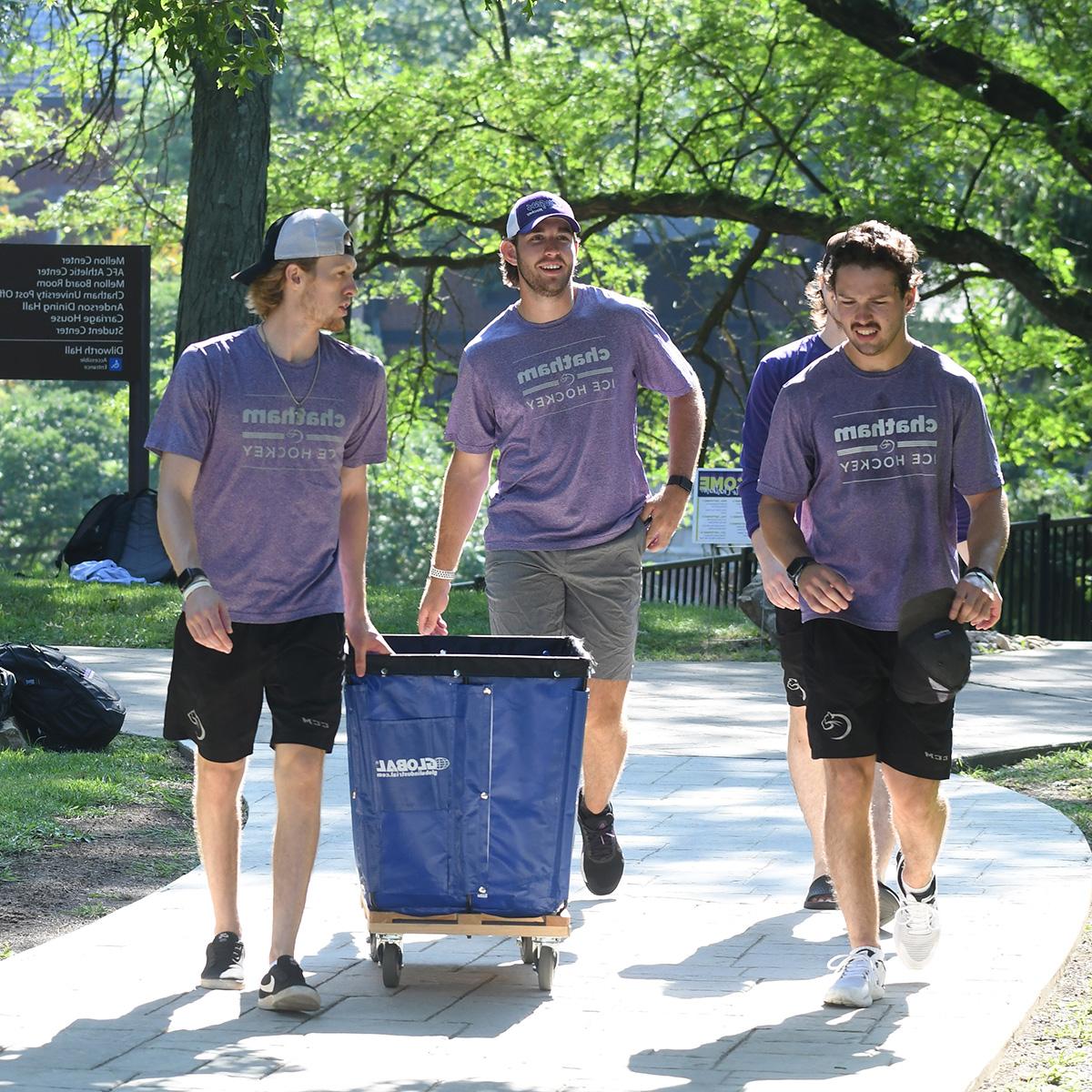 Photo of four young men wearing purple Chatham University Men's Hockey t-shirts and pushing a moving cart