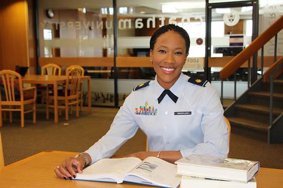 Photo of a woman in military uniform sitting at a library desk, smiling, with a book open in front of her