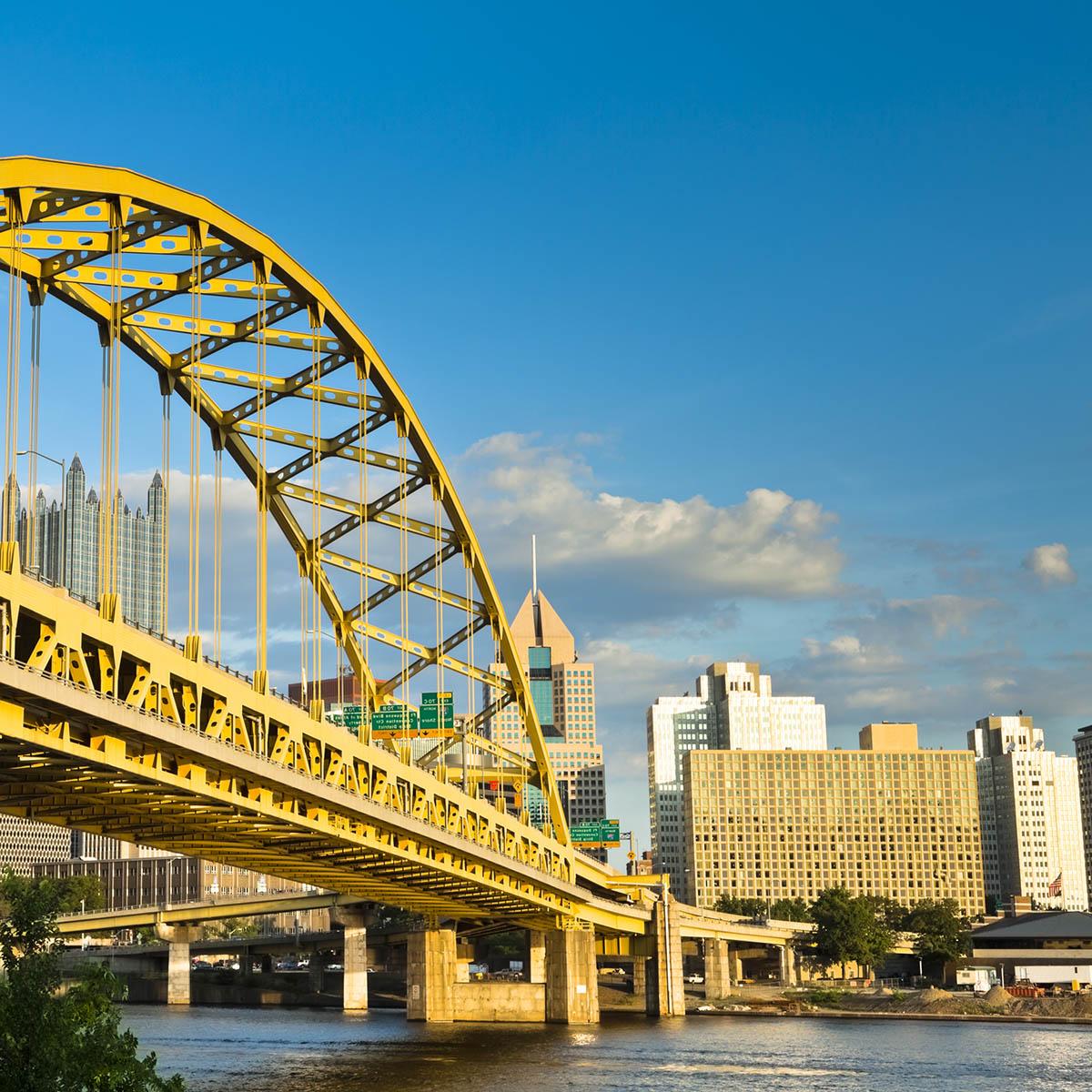 Photo of a yellow bridge over a river and the Pittsburgh skyline