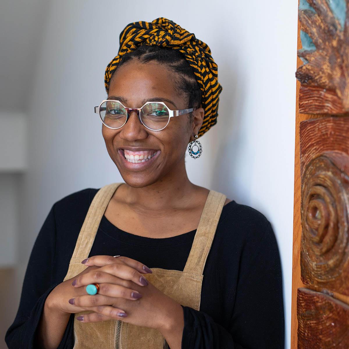 Photo of Ciera Marie Young, a Black woman with hexagonal glasses, smiling in front of a colorful painting
