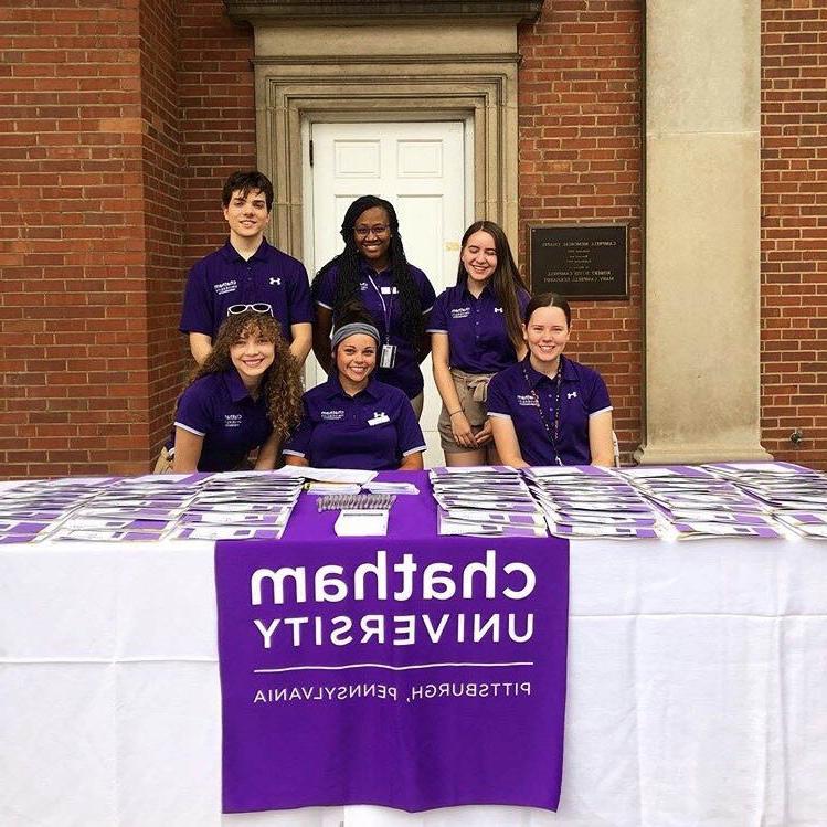 Photo of six Chatham University students in purple shirts, working at a table outside on Shadyside Campus