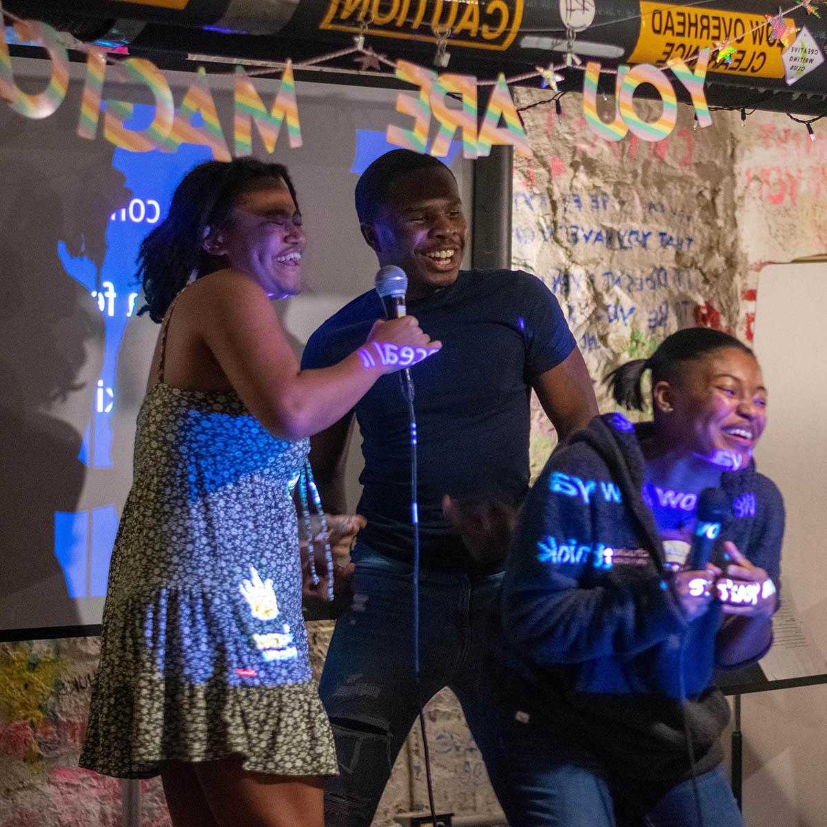 Photo of four 黑色的 students laughing and sining on stage with microphones during BIPOC 卡拉ok night. There is a sign behind them that says YOU ARE MAGIC.